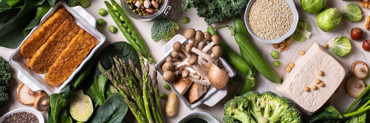 An overhead spread of plant-based foods including tofu, tempeh, mushrooms, asparagus, broccoli, kale, and quinoa